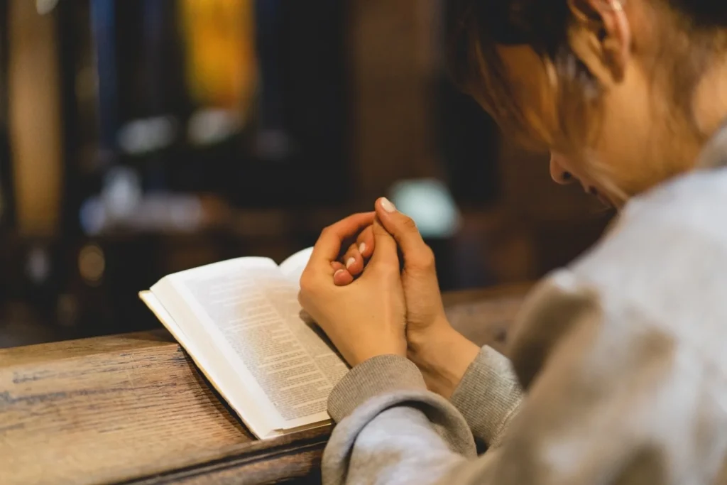 Person sitting at a wooden bench with hands clasped in prayer over an open book, possibly a Bible, in a dimly lit setting.
