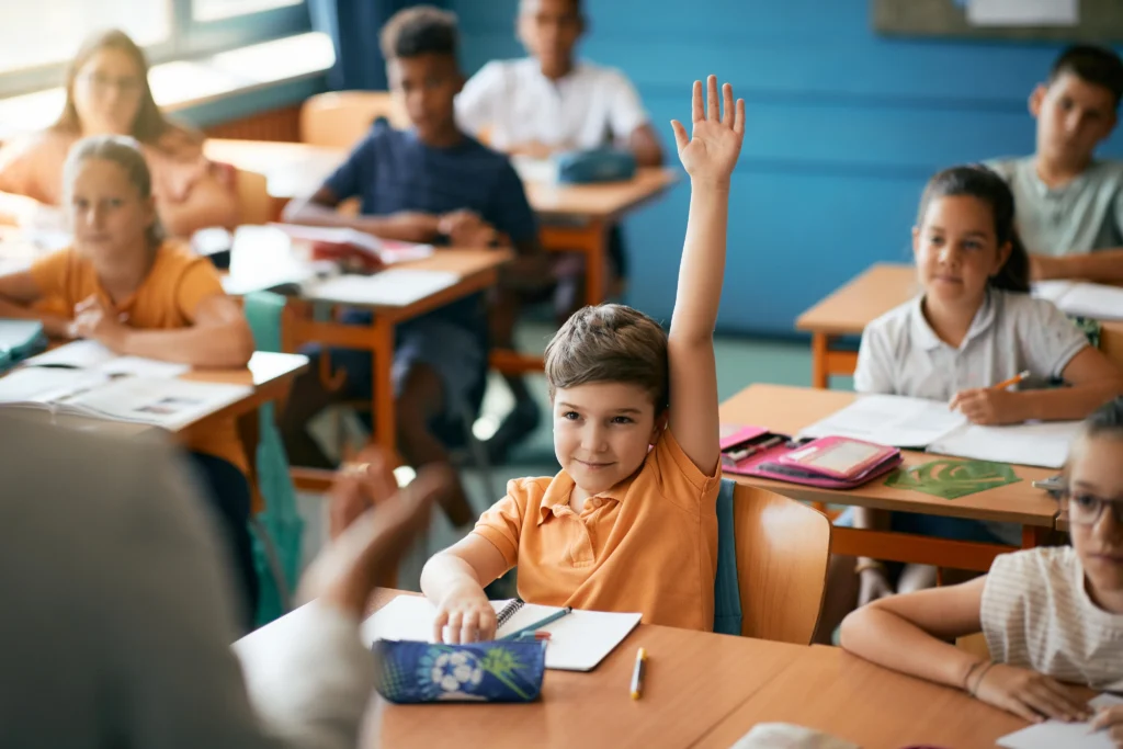 A young student raises his hand in a classroom while others sit at their desks with notebooks and textbooks open, facing the teacher.