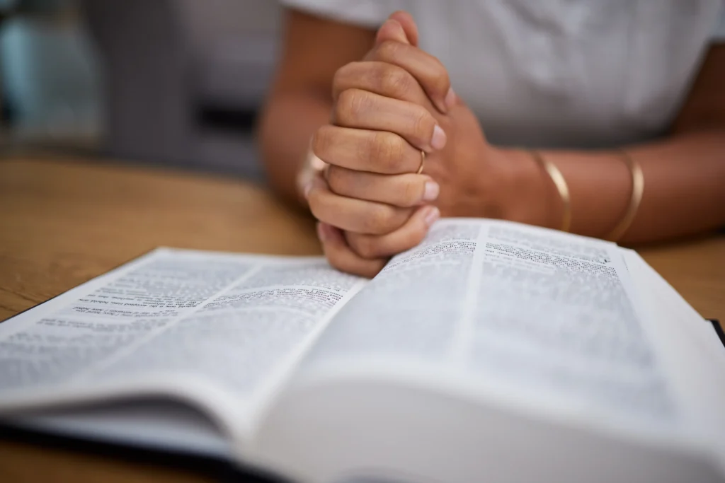A person sits at a table with hands clasped in prayer over an open Bible.