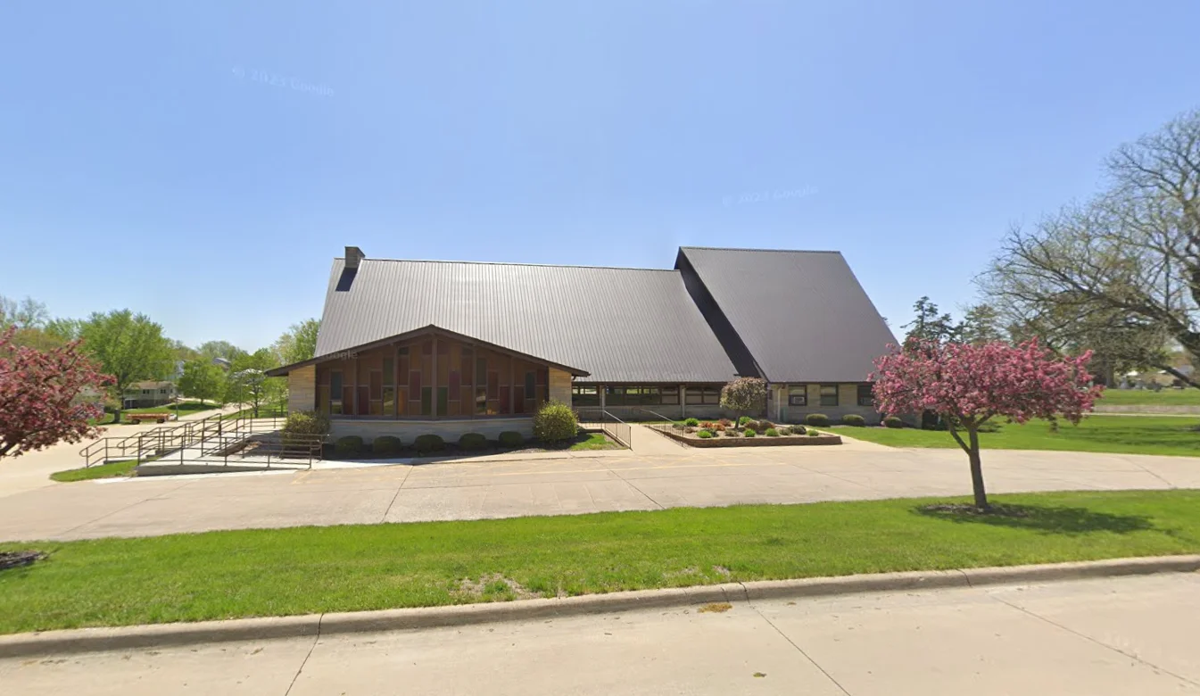 A modern building with a sloped roof, accessible ramp, and landscaped surroundings, set against a clear blue sky with blooming trees in front.