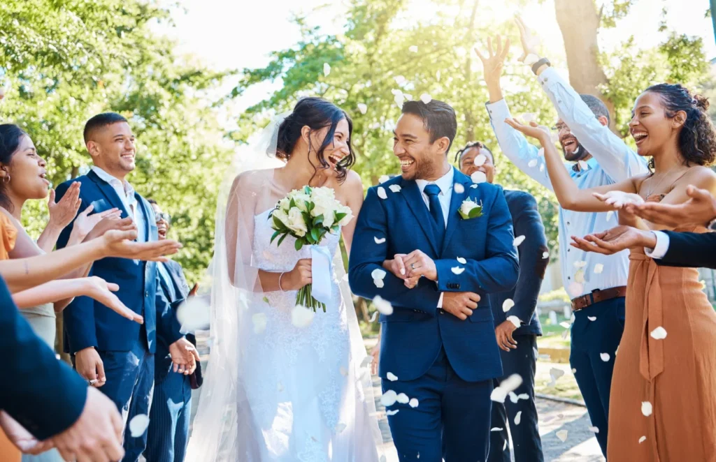 A bride and groom walk outdoors, smiling and holding hands, as guests throw white flower petals in celebration.