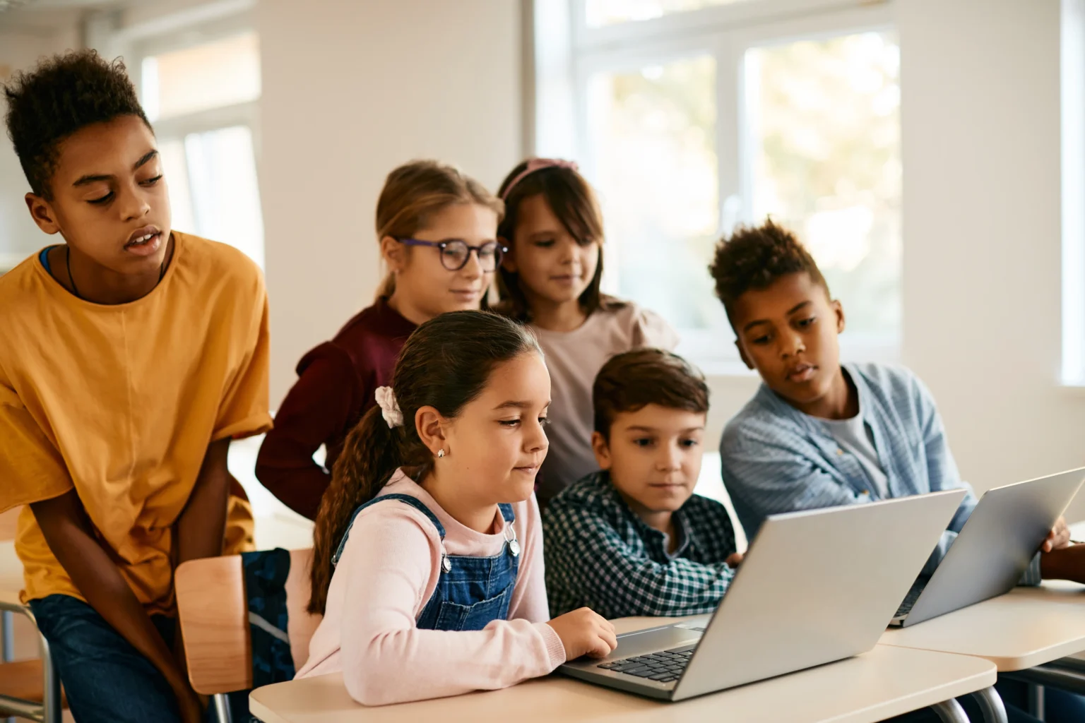 Six children sit and stand around two laptops on a classroom desk, collaborating and looking at the screens in a well-lit room.
